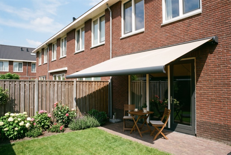 Uitgerold knikarmscherm met zandkleurig doek boven het terras van een bakstenen rijwoning